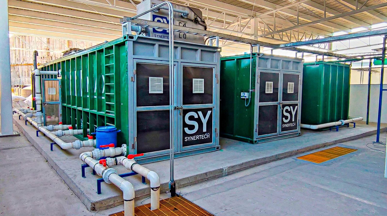 Técnico inspeccionando una planta de tratamiento de aguas residuales modular de color verde marca BIOMAX SYNERTECH, que incluye módulos de pretratamiento, decantación, DAF y oxidación avanzada. La planta está ubicada en un entorno industrial con techo alto y tuberías visibles. - ver más información